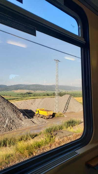 This is a photograph taken from inside a train compartment, looking out of the window at a rural industrial landscape. The scene features a bright yellow dump truck parked on a dirt road next to large piles of gravel. A tall, metallic power line pylon stands prominently in the middle ground. In the background, rolling green hills and fields stretch towards a clear blue sky. The window itself has some reflective text, likely a warning about not opening the window due to air conditioning. The overall impression is one of a sunny day in an industrial or construction area located in a rural setting. The specific location of Spišský Štiavnik, Slovakia, is relevant context for this scene, suggesting a geographical setting within that region.