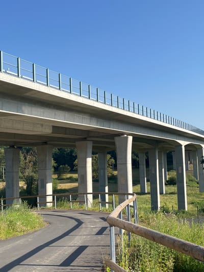 The image shows a modern concrete overpass with a green metal railing against a clear blue sky. The overpass is supported by numerous concrete pillars. In the foreground, a paved pathway curves to the left, bordered by a wooden railing and lush green grass with some wildflowers. The environment suggests a rural or semi-rural setting. The location is identified as Momberg, Germany.