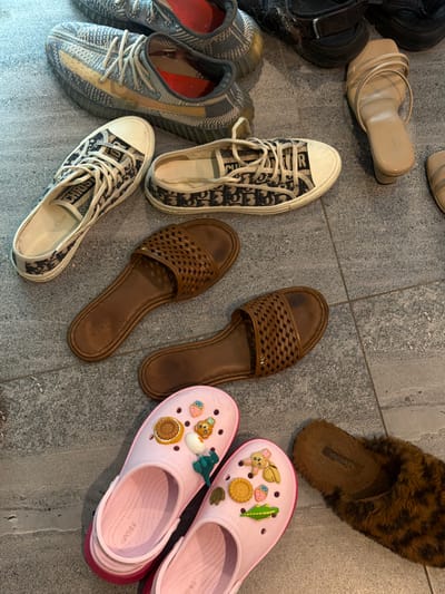The image depicts an assortment of footwear arranged on a gray tiled floor, likely within an indoor space. The footwear includes: two pairs of sneakers with blue and beige patterned exteriors and white laces; a pair of gray and beige Adidas Yeezy sneakers; a pair of tan leather sandals; two brown woven leather sandals; a pair of brown faux-fur lined slippers; and a pair of pink Crocs adorned with Jibbitz charms. The location is not discernable as Kerobokan, Indonesia, based solely on the visual information provided. There are no individuals visible in the scene, and the only activity implied is the placement of the shoes on the floor.