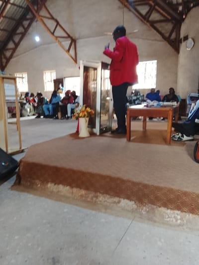 The image shows a person wearing a red jacket standing on a stage speaking to a group of people sitting on chairs and at a table. There is a flower arrangement in a white vase on the stage. The scene is indoors and the walls are beige. The floor is made of concrete and has a brown carpet on the stage. The location is a room in a building in the city of Mukaiya, Nigeria, based on the provided coordinates.