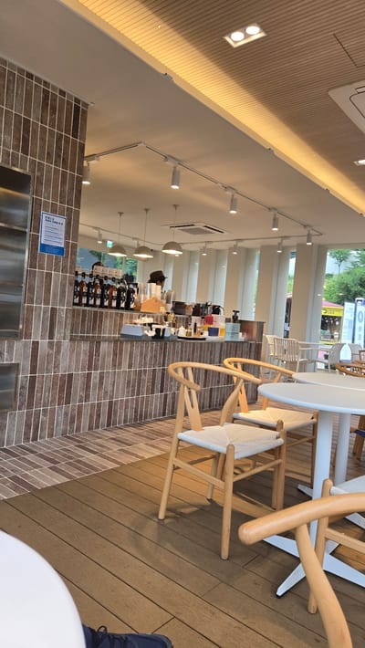 The media file shows the interior of a modern cafe with wooden flooring and brick walls. Several wooden chairs with woven seats are placed around white tables, suggesting a seating area for customers. In the background, a coffee bar is visible with various bottles of syrups and coffee-making equipment. A person wearing a hat and a dark top can be seen behind the counter, possibly a barista. The cafe appears to be well-lit with track lighting and recessed ceiling lights. A sign with Korean text is posted on the brick wall near the seating area. Outside the cafe, through large windows, a street scene is partially visible with trees and buildings, indicating an urban or suburban location. The overall ambiance of the cafe seems relaxed and contemporary. The time of day appears to be daytime, as evidenced by the natural light coming from the windows and the lighting within the cafe. There are no clear indicators of the weather, but the interior lighting suggests it might be overcast or late afternoon. The presence of a coffee bar and seating arrangement clearly points to the setting being a cafe.