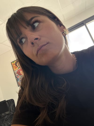 A person with brown hair and brown eyes is looking up at the camera. They have long eyelashes and are wearing a gold earring. A picture of candy wrappers is in the background, and a window is in the upper left corner of the frame. Part of an office chair is visible on the lower right. The setting appears to be indoors, possibly an office. There is no indication of the city of Les Arcs, France.