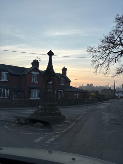 The image depicts a road junction in Ruyton-XI-Towns, United Kingdom, at either dawn or dusk. In the immediate foreground, a dark, weathered stone market cross stands on a small, circular, rock-lined traffic island. The cross features a tiered octagonal base, a central shaft with an arched niche containing vertical bars on one face, and a finial at its apex.
Behind the monument, to the left, are several two-story red-brick buildings with dark-tiled roofs and multiple white-framed sash windows. An antenna is visible on a chimney stack of the leftmost building. Overhead, multiple horizontal utility lines traverse the upper portion of the frame. To the right of the cross, a large deciduous tree, currently bare of leaves, presents a dark, intricate silhouette against the sky.
The asphalt road, which appears damp or reflective, encircles the monument and extends into the background, displaying faded white road markings. In the distant background, a large stone structure, possibly a church or castle, is visible atop a hill, partially obscured by atmospheric haze. The sky transitions from a pale blue overhead to a soft orange and yellow hue near the horizon. The vantage point is from inside a vehicle, with a portion of the dashboard visible along the bottom edge of the image. No human figures or active vehicles are present in the scene.