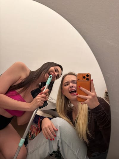 Two women strike playful poses in a selfie taken in front of a round mirror. One woman, wearing a pink top and holding a mop handle like a microphone, sings with exaggerated emotion. The other woman, with blonde hair and dressed in a brown shirt and jeans, takes the picture with a phone, mimicking the singing pose. The reflection captures a casual indoor setting with light walls, creating a fun and lighthearted atmosphere.