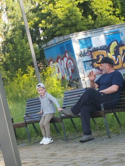 In Kingisepp, Russia, on a sunny day, a man and a boy are seated on a park bench. The man, wearing a dark blue t-shirt and a black baseball cap, is on the right side of the bench. His left leg is crossed over his right, and he is gesturing with his right hand. The boy, on the left side of the bench, is wearing a light grey long-sleeved shirt, tan pants, a white baseball cap, and white and grey sneakers. He is looking towards the camera. Behind them, a blue structure with graffiti is visible, along with lush green trees and foliage. The ground is paved with light-colored bricks.