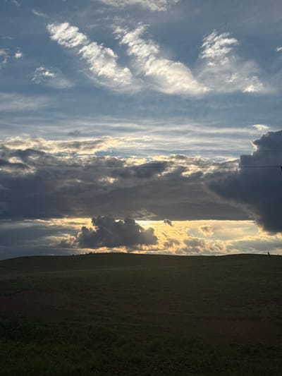 The image shows a field of green grass taking up the right side of the frame. To the left of the field, a cloudy sky is visible. The clouds range in color from white to dark gray. Sunlight peeks through the clouds in the center of the frame, creating a bright spot. Power lines stretch across the sky near the center. The image is rotated 90 degrees clockwise. There is no evidence to suggest this image was taken in Batmany, Russia.