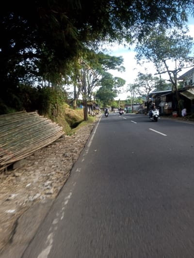 The scene depicts a daylight view of a two-lane asphalt road in Phonengam, Laos, observed from a low angle, possibly from a moving vehicle. The road features a solid white line marking the right edge and intermittent white dashed lines in the center.

On the immediate left, a dirt and gravel shoulder runs alongside the road, leading to a shallow ditch. Beyond the ditch, a steep, vegetated embankment covered in green foliage rises. Resting on this embankment, parallel to the road, is a large, ordered stack of cut bamboo poles. The left side of the frame is heavily shaded by dense overhead tree canopy, likely bamboo, obscuring the sky.

On the right side of the road, the shoulder is narrower, transitioning to an unpaved area. Utility poles with overhead lines are visible along this side, situated in front of a series of single-story structures with corrugated metal roofs and various lean-tos, consistent with roadside shops or dwellings. Further down, more substantial buildings are visible.

Several individuals are operating motorcycles on the road. In the foreground on the right, a person in a light-colored shirt is riding a white scooter, traveling away from the camera. Further down the road, multiple other motorcycles, primarily dark-colored, are also moving in the same direction, indicating active traffic flow. The sky above the right side of the road is partially visible, showing a clear blue sky with some scattered white clouds. The overall environment suggests a semi-rural or peri-urban setting with commercial and residential elements integrated into the natural landscape.