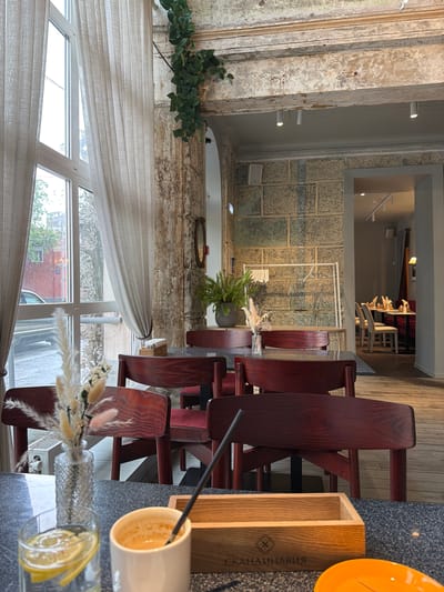 The image shows the interior of a café in Перовское сельское поселение, Ukraine. The café features rustic, textured walls and a large window with a sheer curtain. In the foreground, there's a table set with a glass of water with a lemon slice, a coffee cup, and a wooden container. The table is surrounded by dark red wooden chairs. There is a vase with dried flowers on the table. A plant in a pot sits further back on the table. Through a rectangular opening, another area of the café can be seen with additional tables and chairs.