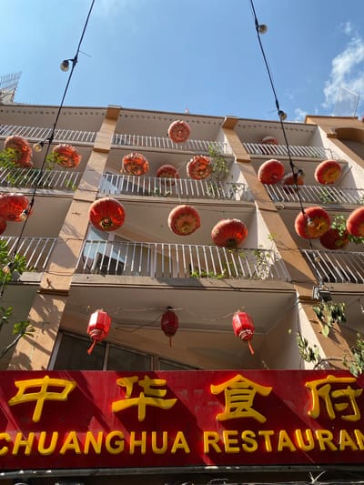 The image depicts the facade of a building adorned with numerous red Chinese lanterns, suggesting a festive or cultural event. The building appears to be a restaurant, with a prominent red sign featuring yellow Chinese characters and the English words "CHUANGHUA RESTAURANT" in yellow lettering. The building has multiple levels with balconies, each lined with white railings. Some balconies have sparse greenery. The lanterns are suspended from wires strung across the building and the street, illuminated by what appear to be bare light bulbs. The sky above is clear blue with a few wispy clouds. The architectural style and the presence of Chinese lanterns strongly suggest the location is within Kathmandu's Thamel district, a known hub for international tourism and diverse cultural establishments, including Chinese restaurants. No people are visible in the image.