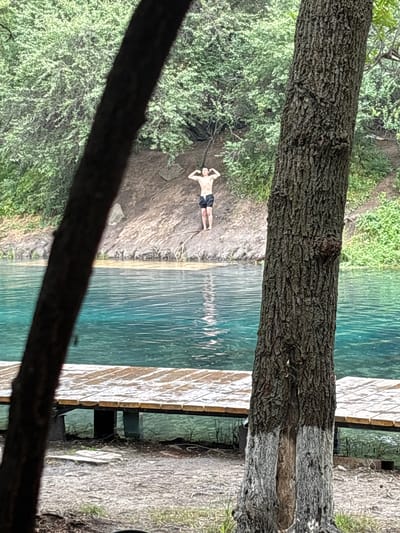 In the city of Zhasqueshu, Kazakhstan, a young man is pictured standing on a rocky bank next to a clear turquoise body of water. The man, wearing black swim trunks, poses with his hands on his hips, flexing his biceps. He stands in the center of the frame, with lush green trees forming a dense backdrop behind him. In the foreground, a wooden pier extends into the water. The scene is framed by the trunks of two trees, one on the left and one on the right, their rough bark contrasting with the smooth water and the man's skin. The overall impression is one of a serene natural setting, perhaps a popular spot for swimming or recreation.