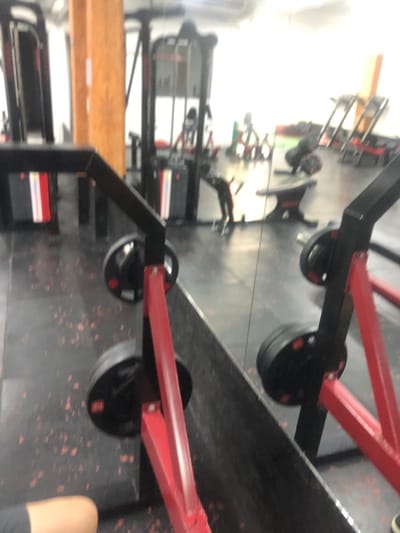 The image displays the interior of a gymnasium located in San Isidro, Argentina. The perspective is low, looking across a dark, speckled rubber floor towards various exercise equipment. A large mirror, reflecting portions of the gym, bisects the scene horizontally in the mid-ground.
In the immediate foreground, the lower left quadrant shows the bare forearm and upper thigh of an individual, dressed in dark shorts, indicating the photographer's position. Adjacent to this, partially obscured by the mirror, are elements of a red and black weightlifting machine, including its red metal frame and black circular weight plates.
The background, visible both directly and as a reflection in the mirror, features multiple pieces of gym equipment. These include cable machines, weight racks, and exercise benches, predominantly black with accents of red, green, and silver. A tall, light-brown wooden pillar stands on the left side of the frame. Within the mirror's reflection, a blurry figure of a person in dark clothing is discernible in the mid-ground, appearing to be bending over or interacting with equipment. The area is brightly lit, resulting in some overexposure in the distant background.