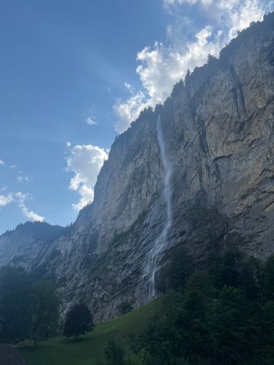 The image displays a prominent waterfall cascading down a colossal, steep rock face in Lauterbrunnen, Switzerland, likely Staubbach Falls. The cliff, predominantly dark grey with visible strata and fissures, rises vertically and extends across the entire right side of the frame. Green coniferous trees line the upper edge of the cliff.
The sky occupies the upper-left quadrant, featuring a clear blue expanse dotted with scattered white cumulus clouds. Rays of sunlight (crepuscular rays) are visible emanating downwards from a cloud in the upper-left.
At the base of the cliff, a verdant grassy slope is visible, interspersed with a few deciduous trees. Denser green foliage, consisting of various trees and bushes, is present in the lower-right foreground and along parts of the cliff's lower sections. A small portion of a reddish-brown tiled roof of a structure is visible in the extreme lower-left corner of the frame. No individuals or animals are present. The scene is illuminated by bright daylight.