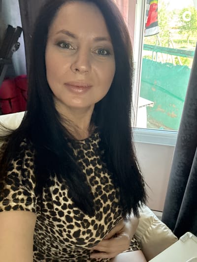 A woman is pictured in a close-up, rotated shot. She has dark hair and is wearing a leopard-print top. She is looking towards the camera with a slight smile. In the background, a window is visible, with some greenery and what appears to be a structure outside. A pillow is also visible behind her. The lighting suggests it is daytime.