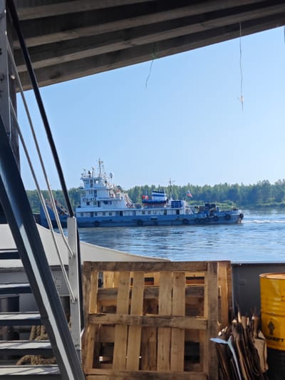 This is a medium shot, taken from a low angle, of a canal or river in Samkovo, Russia. The foreground is dominated by the deck of a vessel, with wooden pallets and a yellow barrel visible on the right. A portion of a ship's stairs is also on the left. In the mid-ground, a large white and blue ship with the identification "OTA-872" is moving down the waterway. The ship has several decks and a Russian flag flying. The background consists of a dense green forest lining the opposite bank of the river, under a clear blue sky. The lighting suggests it is daytime, with sunlight illuminating the scene. There are no people visible in the image.