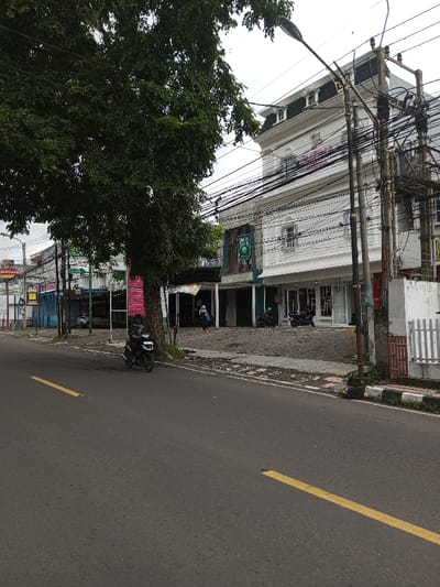 The image displays an urban street scene in Vaatharawattai, Sri Lanka, under an overcast sky. A dark asphalt road with two broken yellow lines spans the foreground. On the left, a large, densely foliated green tree dominates, partially obscuring the background. A single individual, dressed in dark clothing, is seated on a black motorcycle positioned on the road near the tree line. On the right, a raised, grey-paved sidewalk runs parallel to the road, fronting a series of multi-story commercial buildings. The most prominent structure is white, featuring architectural details, a mansard roof, and a pink-and-purple sign on its upper facade. Adjacent to this building is a darker-facade building displaying a green sign with white text. Multiple utility poles, heavily laden with a dense network of black electrical and communication wires, are prominent along the right side of the street. Two individuals are visible on the sidewalk in front of the darker-facade building; one appears to be wearing a head covering. Two additional motorcycles are parked on the paved area. A white picket fence segment and a low-level structure with a reddish-brown brick pattern are present on the far right.