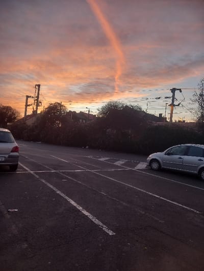 The image captures an outdoor scene at sunrise or sunset in Sallaumines, France. The upper sky displays vibrant orange, pink, and light blue clouds, bisected by a distinct, elongated, reddish-orange streak extending diagonally from upper left towards the lower mid-right. The foreground consists of a dark asphalt surface with faded white parking or road markings. A silver hatchback's rear quarter is visible on the far left. Another silver hatchback is parked facing left on the right side of the asphalt. Minor debris is scattered on the ground. The mid-ground features a dense line of dark green vegetation. Behind this vegetation, residential or commercial buildings are partially visible. Two tall metal utility poles with visible wires stand against the sky on both the left and right sides of the background, with horizontal lines stretching across the scene.