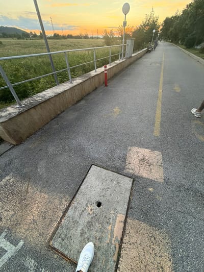 The image depicts a paved pathway in the city of Pravets, Bulgaria, with a railing separating it from a grassy field. The pathway is marked with a yellow line and has a manhole cover in the foreground. In the background, there are trees, power lines, and a sunset sky. A person is visible on the pathway, walking away from the camera.