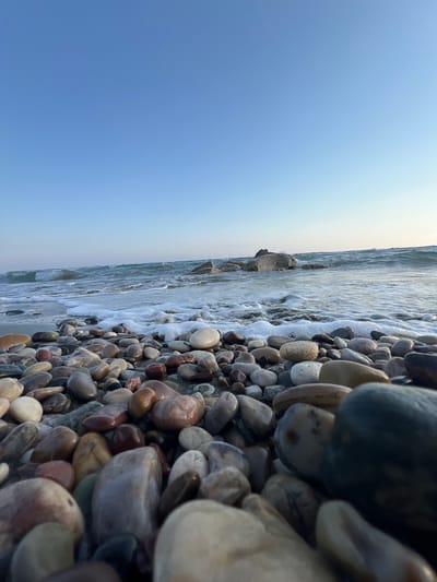 This is a close-up, low-angle, sideways photograph of a pebbly beach at Episkopi, Cyprus. The frame is divided into two main sections: the shore and the sky.

The shore on the left is densely packed with smooth, rounded pebbles of various sizes, shapes, and colors, including shades of gray, brown, white, red, and tan. Some pebbles are wet and appear darker, reflecting the light. The water is gently lapping at the shore, creating frothy white waves and foam that wash over some of the pebbles. In the distance, larger rocks are visible, and further out, the sea is a calm, blue expanse.

The right side of the image is dominated by a clear, pale blue sky, suggesting a bright, possibly sunny day. The transition from the horizon where the sky meets the sea is subtle, with a slight haze or lighter tone near the water's edge.

The perspective is from ground level, looking across the pebbles and the water towards the horizon and sky. The close-up view of the pebbles emphasizes their texture and variety. The overall mood of the image is peaceful and serene, capturing the natural beauty of a beach. The time of day appears to be late afternoon or early evening, given the soft light and the angle of the sun, casting a gentle glow. No people or text are visible in the image.