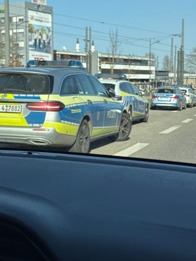 The image, taken from within a vehicle looking forward, displays a line of five German police vehicles on a multi-lane asphalt road in Kehl, Germany, under clear daylight conditions.
In the mid-ground, a silver/grey Mercedes-Benz E-Class Estate (S213 generation) police vehicle is positioned nearest to the viewer. It features blue and yellow striped livery, "POLIZEI" text on its side, and blue emergency lights mounted on the roof. The rear license plate is partially visible, showing "OG 4 7882". Directly behind this vehicle is another police station wagon, similarly marked with blue and yellow livery and equipped with blue roof lights. Further down the road, at least three more police vehicles, appearing to be BMW sedans, are visible in the same blue and yellow livery with blue roof lights. All vehicles are aligned in a single lane, indicating they are stopped or moving slowly in a procession. No occupants or individuals are clearly visible interacting with the vehicles.
The asphalt road features white dashed and solid lane markings.
In the background, on the left side, a multi-story building displays a large advertising banner depicting people and partial text including "Troppeau". Two prominent, pointed architectural structures, identified as minarets of the Kehl Mosque, are visible against a clear blue sky between urban buildings. The background also includes additional buildings, bare deciduous trees, and overhead utility lines with supporting poles. The foreground consists of the dark blue dashboard and upper window line of the camera-operating vehicle.