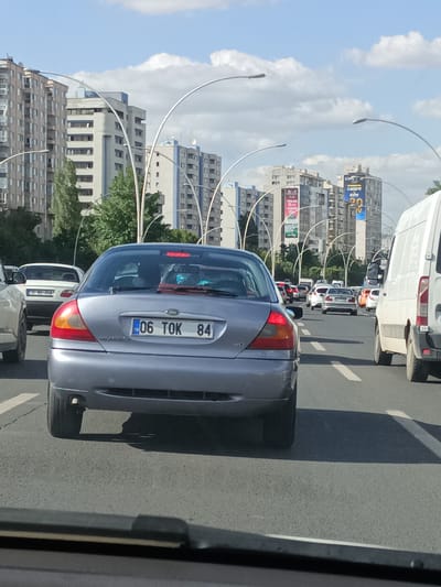 A silver Ford Mondeo is captured from behind, in moderate traffic on a multi-lane road. The car's license plate reads "06 TOK 84". The scene is set in a city environment with tall, modern apartment buildings lining the sides of the road. Lampposts with curved arms are visible along the median. The weather appears to be clear with some clouds in the sky, suggesting it is daytime. The overall atmosphere is one of urban movement and daily commuting. The perspective is from within another vehicle, looking forward. The background shows a continuation of the road with other vehicles, including a white van to the right and other cars further ahead. Advertisements are visible on some of the buildings. The location cues, such as the architecture and the license plate format, point to Yenimahalle, Turkey.