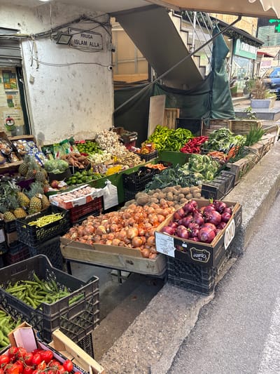 Here is a detailed description of the image:
**Location:** The image appears to depict a street market scene in Tirana, Albania, evidenced by a sign reading "Rruga Islam Alla," which translates to "Islam Alla Street."
**Scene:** The photograph shows a variety of fresh produce displayed for sale along the side of a street. A collection of fruits and vegetables are arranged in crates and on shelves, including red onions, brown onions, potatoes, carrots, green peppers, red peppers, green beans, tomatoes, pineapples, apples, garlic, limes, and other unidentified vegetables.
**Environment:** The produce stand is situated along a sidewalk next to a street. A building with a storefront and awning is visible behind the produce displays. The street surface is concrete. The general lighting conditions suggest it is daytime.