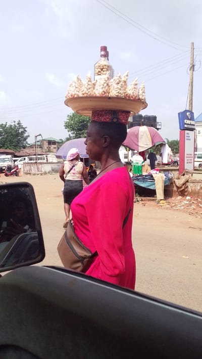 The image depicts a daytime street scene in Igbo Ukwu, Nigeria, observed from a low vantage point, possibly from a vehicle. The primary subject is a dark-skinned woman in profile, positioned in the mid-ground, facing left. She wears a long-sleeved, bright pink dress and a brown shoulder bag. On her head, she expertly balances a light-colored, shallow tray, which holds a clear glass bottle filled with peanuts, surrounded by multiple small, clear plastic bags also containing peanuts, arranged in a conical display. A patterned fabric ring rests on her head beneath the tray.

In the background, a second woman walks away from the viewer, clad in a light-colored top, dark skirt, and a light head covering. Further down the street, several market stalls with large, colorful umbrellas are visible, under which indistinct figures are present, indicative of commercial activity. To the right, a blue and white sign for "CAP 10" advertises "FUEL KERO DIESEL," "MINI MART," and "CAR WASH." A wooden utility pole with overhead power lines stands near the sign. Various buildings, including multi-story structures and one with water tanks on its roof, are visible in the distant background. On the left side of the unpaved or dirt road, a white van/bus and a red motorcycle are present. Debris and scattered goods are visible on the ground near the market area.