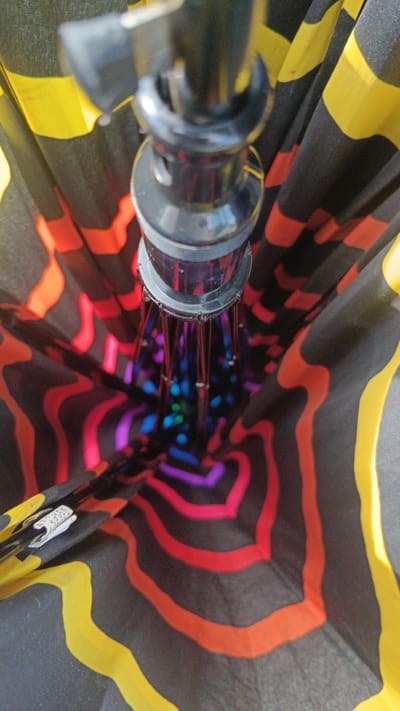 The image provides an extreme close-up, low-angle view looking up into an open umbrella. The umbrella fabric features a vibrant, wavy pattern of black, yellow, red, orange, purple, and blue. The umbrella's internal mechanism, a black pole with metallic struts, is visible at the top, extending down into the fabric. The overall impression is one of abstract color and geometric design, with the light catching the metallic elements and the fabric's saturation. The image is set in Sankt Pölten, Austria, but no specific landmarks or environmental context beyond the umbrella itself are identifiable. The scene is devoid of people or any discernible action.
