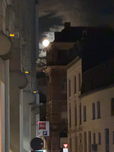 This is a nighttime street scene in Ghent, Belgium, featuring the moon visible through clouds, illuminating the facades of buildings. On the left, a lighter-colored building with curved architectural elements is partially visible, featuring two orange rectangular lights. To the right of this, a darker brick building with a distinct window structure is visible, and further to the right, a lighter-colored building with multiple tall, narrow windows. In the foreground, on a pole, there is a sign with the "ibis" logo in red and white, accompanied by a blue parking sign with a white "P" and a white arrow pointing left. Below this, a black circular object is mounted on a pole. Further down the street, another traffic sign with a red-bordered white triangle pointing downwards is visible. The scene is dimly lit by the moonlight and artificial lights from the street and buildings.