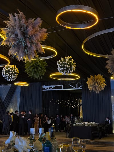 The image depicts an indoor event space, likely a reception hall, decorated with large, circular, illuminated fixtures and hanging floral arrangements. In the foreground, a table is set with glassware and bottles. The middle ground shows a gathering of people, many dressed in formal attire, some interacting in groups. A long, black-draped table is set for a formal meal in the background, with chairs arranged around it. The overall atmosphere suggests a special occasion such as a wedding reception or formal party. The location is identified as San Francisco Ocotlán (Ocotlán), Mexico.