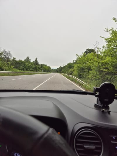 The image is taken from the perspective of a driver inside a car, looking out at a two-lane road lined with trees. The road appears to be in a rural or semi-rural area, with a guardrail on the left side and dense green foliage on both sides. The sky is overcast and a light gray. In the foreground, the car's dashboard is visible, including a black car phone mount attached to the windshield, part of the steering wheel, and an air vent. There is a digital display on the dashboard showing "15 70x". The location is identified as a road in the vicinity of Ponor, Bulgaria.