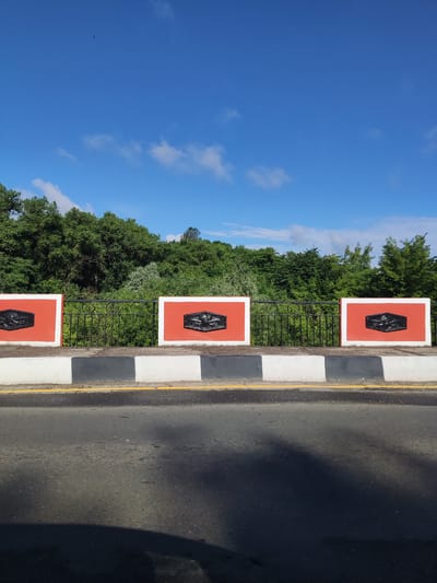 This media file depicts a view of a road with a decorative barrier and a lush green treeline against a bright blue sky with scattered white clouds. The barrier consists of three rectangular panels, each with an orange background and a white border. Centered on each orange panel is a black, hexagonal emblem with intricate detailing. A black wrought-iron fence runs along the top of the barrier. Below the barrier, there is a road with distinct white and grey markings, and a thin yellow line bordering the edge of the asphalt. The treeline behind the barrier is dense and appears to be in full foliage, suggesting a warm season. The sky is clear and bright, indicating it is daytime, likely late morning or early afternoon, with fair weather. There are no people or specific activities visible, presenting a static, scenic view. The overall impression is one of a well-maintained roadside with an artistic flourish, set against a natural backdrop. The location context suggests this scene is in Polatsk, Belarus. No visible text is present in the image.