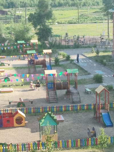The image depicts a children's playground located in Askiz, Russia. The playground features various structures including slides, swings, a seesaw, and climbing equipment, all set on a sandy surface. Colorful flags are strung across parts of the playground, adding to the festive atmosphere. Several children are visible playing on the equipment, and an adult appears to be supervising. In the background, a grassy field and trees can be seen, suggesting a semi-rural or suburban setting. The playground is enclosed by a low, multi-colored fence. The overall scene captures a typical day at a community playground.