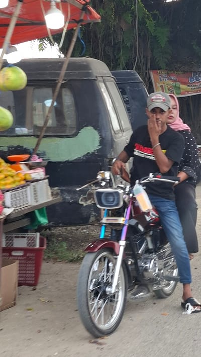 In Keudebayu, Indonesia, a person wearing a gray cap and a black t-shirt is sitting on a motorcycle, with a person wearing a pink hijab behind them. The motorcycle has a silver and chrome engine, a maroon colored body, and a colorful exhaust pipe. The driver's left hand is raised towards their face. They are wearing blue jeans. The person in the back is wearing a gray dress with black polka dots. The scene is in front of a black van, with a red canopy above. To the left of the van, there are fruits displayed for sale, including coconuts and oranges, with scales and crates. A sign with text in Indonesian is visible in the background. The ground is paved with asphalt and dirt.