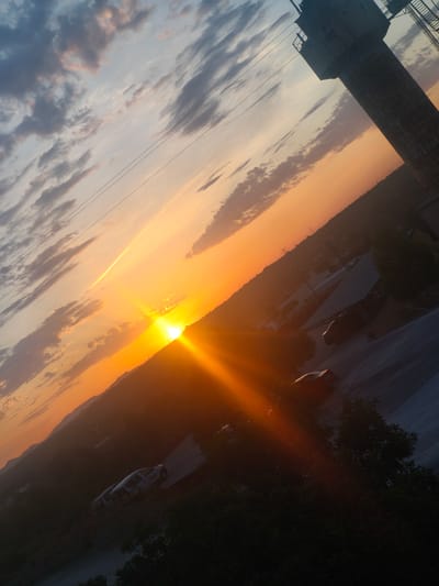 The image captures a dramatic sunset over Riba-roja de Túria, Spain. The sky is a vibrant canvas of orange and yellow hues, with scattered clouds tinged pink and gray. A tall, industrial-looking tower, possibly a water tower or communication tower, stands silhouetted against the sky on the right side of the frame. In the foreground, a road slopes downward, with several vehicles visible. One white van is parked on the road, and another darker car is further down. The landscape is characterized by rolling hills and scattered trees, with some buildings and structures visible in the distance. The perspective is angled, giving a sense of looking down onto the scene. Power lines are also visible crossing the sky.