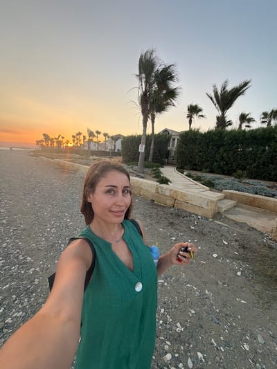 An adult female is taking a selfie on a pebbly beach at sunset in Pervolia, Cyprus. The woman is wearing a green sleeveless dress with a V-neck and a white button. She has medium-length brown hair and is wearing a silver necklace. In her right hand, she is holding car keys and a blue object, possibly a drink. The background shows palm trees, a hedge, a walkway made of stone blocks, and some buildings. The sun is setting over the sea, casting an orange glow on the sky and illuminating the palm trees. The beach is covered in small pebbles and sand.