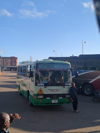 The image shows a green and white bus parked in a bus station in Morogoro, Tanzania. The bus is labeled "Myulingo Express" and has the words "MORO" and "DOM" on the front. There are two people standing near the bus, one in a black shirt and the other in a blue shirt. There is a third person sitting in a wheelchair. The bus is parked on a paved surface and there are other buses parked behind it. There are buildings in the background, and the sky is a clear blue. The scene is taken from a slightly low angle.
