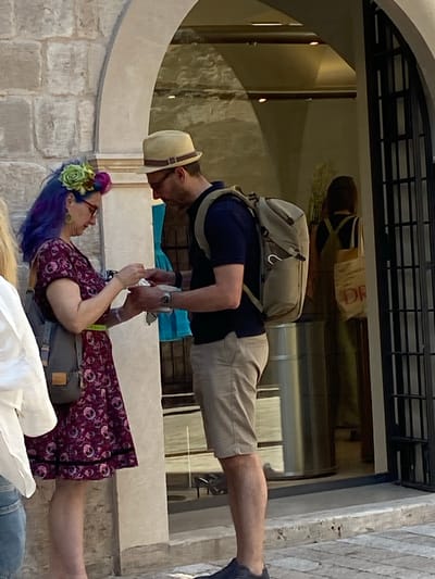 In Dubrovnik, Croatia, a woman with vibrant purple hair adorned with a green and pink floral crown and large circular earrings, wearing a dark patterned dress and a grey backpack, stands facing a man. The man, wearing a light-colored fedora, glasses, a dark blue polo shirt, and beige shorts, is handing the woman a white object. He carries a beige backpack. They are positioned beneath an arched stone structure, possibly an entryway, with a shop visible through an open doorway in the background. Another person is partially visible on the far left wearing a white shirt. The scene is captured during daylight with sunlight casting shadows on the stone pavement.
