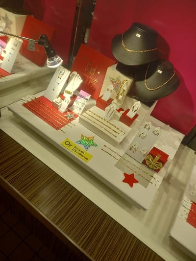 This image captures a jewelry display within a retail setting in Vendin-le-Vieil, France. The central feature is a white display platform showcasing various gold-colored jewelry items, including multiple necklaces, bracelets, earrings, and rings, presented on an assortment of white stands and forms. Two black, neck-shaped busts are positioned in the upper right quadrant, each exhibiting a gold-colored necklace and bearing a small white price tag. A directional spotlight with a silver-colored casing and a clear, faceted lens extends from the upper left, directly illuminating the display. The background consists of a solid magenta-pink wall. Decorative elements on the display include several red cardboard stars, a holographic star sticker, and a small red gift box adorned with a gold ribbon. A yellow label prominently displayed on the white platform reads "Or Garantie 375 millièmes" (Gold Guaranteed 375 thousandths). The immediate foreground consists of a dark brown counter featuring a distinct, striped wood grain pattern, and a dark tiled floor is partially visible in the bottom-left corner of the frame. No individuals are present in the scene.
