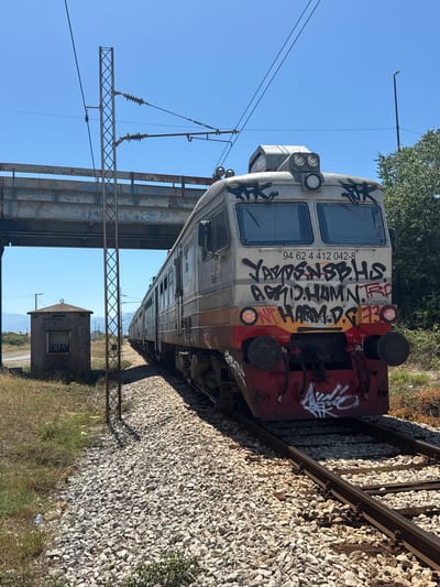 The image depicts a train car on a track in Mojanovići, Montenegro. The train car is heavily graffitied with black and red spray paint. The front of the train car has visible lights and what appear to be brake lines. Text on the train car includes "94 62 4 412 042-8", "YARDENSBHS AGHD.HAMN. TRP", "jeznick prevozne C", and "AD Podgorica". A small, weathered building with the word "KIAH" visible on it is situated near the tracks. Overhead, power lines and supporting structures are visible against a clear blue sky, suggesting a sunny day. The train tracks are surrounded by ballast stones and dry grass, indicating a rural or industrial setting. The overall impression is one of an abandoned or disused train car in a somewhat desolate location.