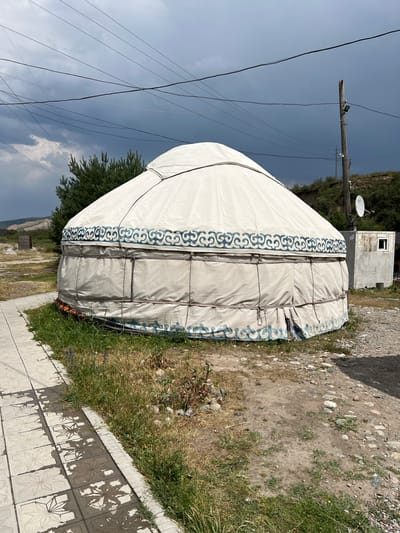 A white yurt with blue ornamental patterns sits on a patch of dry grass next to a tiled walkway. The yurt is circular with a domed roof and appears to be constructed of fabric stretched over a frame. A utility pole with electrical wires and lights stands to the right of the yurt, next to a small, weathered white building with a single window. In the background, a pine tree and a grassy hillside are visible under a dark, cloudy sky. The ground around the yurt is a mix of dirt, gravel, and sparse vegetation. The scene is likely located in Yuryevka, Kyrgyzstan, based on the presence of traditional yurts and the overall landscape. There are no people visible in the image.