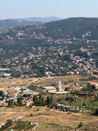 This is a distant, high-angle shot of the city of Mdairej, Lebanon. The image is dominated by a densely populated hillside covered in buildings with red-tiled roofs, interspersed with green trees. In the mid-ground, a large, white statue of Jesus Christ stands on a circular base, overlooking the city. The area around the statue appears to be a religious site, with smaller structures and pathways. The foreground features a dry, grassy landscape with a few scattered buildings and roads. The overall impression is one of a vibrant, lived-in city nestled within a natural, mountainous environment.