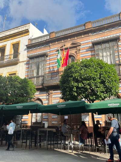 The image captures a street scene in Seville, Spain. It features a building with a facade decorated with alternating stripes of brown and blue tiles, punctuated by ornate arched windows. Three flags, predominantly red, yellow, and green, are visible, hanging from the building's balcony. Below, a restaurant or bar area extends onto the sidewalk, with green umbrellas marked with "Voll-Damm DOBLE MALTA" providing shade to the tables and chairs. People are seated and standing in the outdoor seating area, engaging in various activities. The environment suggests a bustling, tourist-friendly atmosphere in a historic part of the city. The composition of the image is slightly angled, and the scene is well-lit by natural daylight.