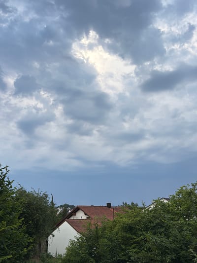 This is a photograph taken outdoors, featuring a view of the sky and a residential area. The image is oriented vertically.
The upper portion of the image is dominated by a sky filled with a mix of dark, dramatic clouds and brighter, sunlit patches, suggesting a partly cloudy or overcast sky. The sunlight breaking through the clouds creates luminous areas.
The lower right portion of the image shows a white building with a red tiled roof, characteristic of residential architecture. This building is partially obscured by lush green trees and foliage that frame the right side of the photograph. The trees are dense, indicating a natural or garden setting surrounding the house. In the distance, other buildings and greenery can be seen, suggesting a suburban or semi-rural location.
The scene appears to be a daytime shot, given the brightness of the sky and the illumination of the clouds. The weather seems to be dynamic, with the presence of both dark and bright clouds. The overall mood is somewhat dramatic due to the cloud formations, but the presence of the houses and trees grounds the scene in a peaceful, everyday setting. There is no visible text in the image. The context provided places this scene in Wettstetten, Germany.