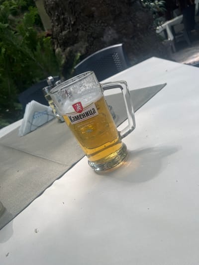 A clear glass mug filled with beer is on a white tablecloth. The mug has a handle and is tilted. The beer is golden in color with foam at the top. There is a label on the mug that reads "Каменица" with a red lion logo above it, and text below that reads "АВТЕНТИЧЕН ВКУС" and "ИСТОРИЯ ОТ 1881". Behind the mug, a stack of white napkins with a blue design is visible, as is a small glass with some liquid. The scene appears to be outdoors at a cafe or restaurant with a large tree trunk in the background, and other tables and chairs are visible in the distance. The location is identified as Bachkovo, Bulgaria.