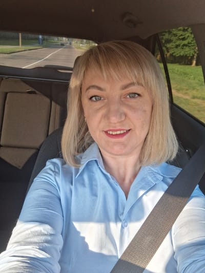 A blonde woman with bangs, wearing a light blue button-up shirt and a seatbelt, is taking a selfie in the driver's seat of a car. The background shows a road with trees and grass, suggesting a location outside of the city center. The woman is smiling and looking at the camera. The image appears to be taken during the day, as evidenced by the natural lighting.