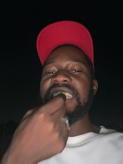 A medium-shot image depicts a Black man with a dark beard and a red baseball cap. He is wearing a white t-shirt and a silver ring on his finger. His right hand is raised towards his mouth, with his fingers slightly inside. The background is dark, and there is no evidence indicating Galadimawa, Nigeria.