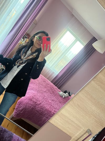 A person with dark hair and glasses is taking a selfie in a mirror. The person is wearing a black blazer, a white shirt, and jeans. Behind the person, there is a bed with a fluffy pink blanket. The background also includes a window with sheer curtains, a desk, and a ceiling light fixture. The room appears to be a bedroom. The mirror is slightly angled, capturing the scene from a tilted perspective.