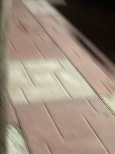 The image displays a severely motion-blurred close-up of a patterned ground surface in Debelt, Bulgaria. The surface consists of alternating horizontal bands of light pink/mauve and off-white/light beige material. Each band features parallel, linear indentations or grooves running diagonally from the upper left to the lower right of the frame. The upper right and far right portions of the image are obscured by deep shadow or darkness. No individuals, animals, distinct objects beyond the patterned ground, or specific actions or interactions are discernible. The scene is characterized by dim and uneven lighting conditions, which, combined with camera movement, results in extreme blur across the entire image.
