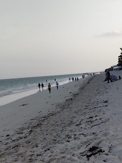 The image displays a sandy beach scene in Gombato Bongwe ward, Kenya, under a light grey sky. The Indian Ocean, characterized by blue-green water and breaking waves, borders the left side of the frame. The beach itself consists of light-colored sand, imprinted with footprints and interspersed with dark organic debris. A minimum of 30 individuals are present across the scene. In the mid-ground, approximately six people are walking along the waterline, with two individuals in the foreground wearing a yellow top and a white top, respectively, and others visible as dark silhouettes. Several individuals are partially submerged in the ocean. Numerous additional figures are distributed along the distant shoreline, predominantly walking. On the far right, two individuals are seated on the higher sand, one wearing a black and white patterned garment, accompanied by a smaller figure. The far right background features a vegetated area with palm trees and indistinct white structures partially visible further down the coastline.