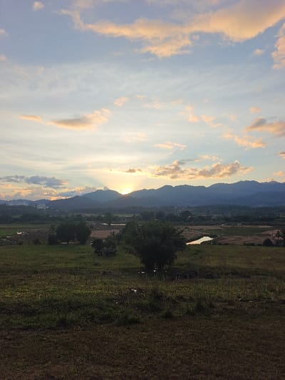 The image captures a rural landscape at sunset in Ban Mae Ping, Thailand. The upper two-thirds of the frame are dominated by a light blue sky with scattered stratocumulus clouds, illuminated by warm orange and yellow light from the setting sun near the horizon. The sun, partially obscured by a range of dark, undulating mountains, is positioned slightly left of center and emits distinct crepuscular rays. The midground consists of expansive fields, primarily green and brown, with several dark green trees. A small, reflective body of water is visible in the mid-right, and a larger irregular patch of saturated ground or water is present in the mid-left. A small, unidentifiable shelter or hut structure is discernible within the fields in the mid-left. The foreground features dark green and brown grassy terrain with visible patches of darker, possibly muddy, ground. No individuals, animals, or active interactions are depicted.